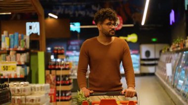 Kahverengi kazaklı Arap bir adam süpermarket koridorunda durup alışveriş arabasıyla alışveriş yaparken gülümsüyor..