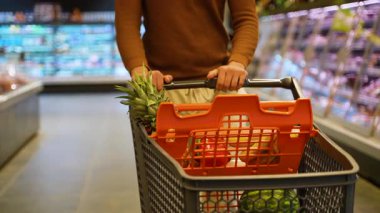 Kahverengi kazaklı bir Arap süpermarketten alışveriş arabası geçirir, market alışverişini seçer ve ziyareti sırasında yiyecek seçeneklerini araştırır..