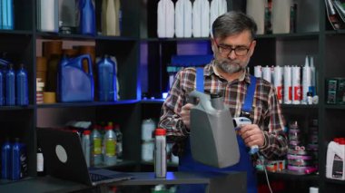 Ekose gömlekli ve tulumlu yaşlı erkek tezgahtar otomotiv envanterini yönetmek için barkod okuyuculu bir motor yağı kabını tarıyor..