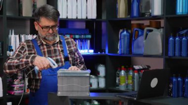 Ekose gömlekli ve tulumlu yaşlı erkek işçi araba aküsünde motor yağı rafları olan bir oto yedek parça mağazasında envanter taraması yapıyor..