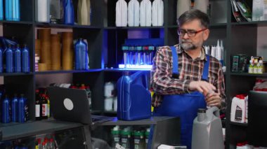 Ekose gömlek ve mavi tulumlu deneyimli bir erkek satıcı bir otomotiv tedarik dükkanında motor yağı konteynırlarını düzenliyor ve kontrol ediyor..