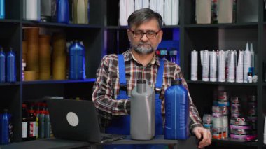 Ekose gömlekli ve mavi tulumlu yaşlı erkek işçi oto yedek parça mağazasında motor yağı kutularıyla tezgahta durup envanter düzenliyor..