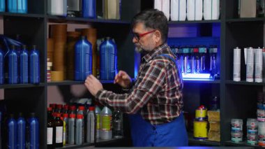 Ekose gömlekli ve tulumlu yaşlı erkek çalışan, envanter kontrolü sırasında bir oto yedek parça mağazasında motor yağı konteynırları düzenliyor..