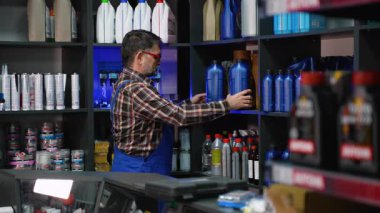 Ekose gömlekli ve mavi tulumlu yaşlı erkek işçi otomotiv parçaları ve malzeme deposundaki bir rafta mavi motor yağı kapları düzenliyor..