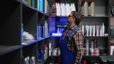 Deneyimli mavi tulumlu ve ekose gömlekli erkek kâtip bir oto yedek parça dükkanında motor yağı raflarını tarıyor ve müşteri için doğru seçeneği seçiyor..