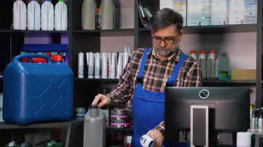 Ekose gömlekli ve tulumlu yetişkin bir katip, bir otomotiv mağazasında envanter sayımı yaparken barkod okuyuculu motor yağı kabını tarıyor..