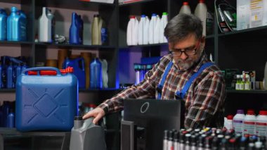 Ekose gömlekli ve tulumlu yetişkin bir katip, bir otomotiv mağazasında envanter sayımı yaparken barkod okuyuculu motor yağı kabını tarıyor..