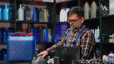 Ekose gömlekli ve tulumlu yaşlı bir erkek, otomotiv parçaları mağazasındaki tezgahta gri bir motor yağı şişesini ve barkod okuyucusunu tarıyor..