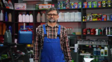 Ekose gömlekli ve mavi tulumlu deneyimli bir erkek tezgahtar, oto yedek parça mağazasında güvenle duruyor ve büyük bir motor yağı kabı sergileniyor..