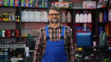 Ekose gömlekli ve mavi tulumlu yaşlı erkek tezgahtar çeşitli motor yağı kapları ve otomotiv ürünleriyle dolu bir oto yedek parça mağazasında duruyor..