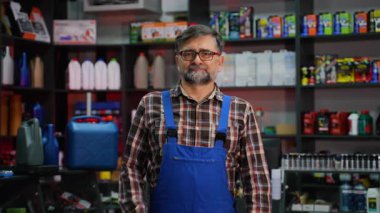 Ekose gömlekli ve mavi tulumlu deneyimli bir erkek satıcı iyi stoklanmış bir oto yedek parça mağazasında tezgahın yanında duruyor..