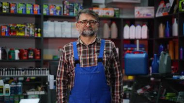 Mavi tulumlu ve ekose gömlekli olgun bir erkek tezgahtar iyi stoklanmış bir oto yedek parça dükkanında yağ, sıvı ve aksesuarlarla poz veriyor..