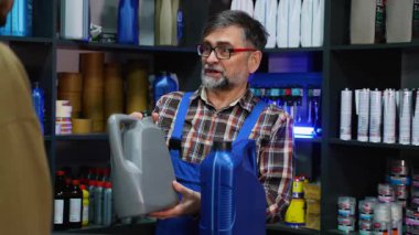 Ekose gömlekli ve mavi tulumlu deneyimli bir erkek kâtip müşterinin motor yağı seçmesine yardımcı olur ve otomobil yedek parça mağazasında ürün detaylarını açıklar..