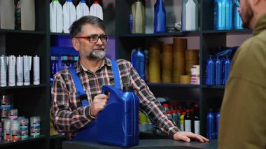 Ekose gömlekli ve mavi tulumlu deneyimli bir katip tezgahta duruyor, büyük mavi bir kamyon yağı kutusunu araba dükkanındaki bir müşteriye teslim ediyor..