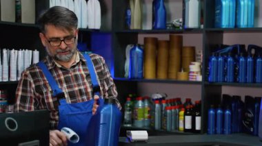 Deneyimli ekose gömlekli ve mavi tulumlu erkek kâtip bir şişe motor yağını tararken, müşteri oto yedek parça mağazasındaki kart terminalini kullanarak ödeme yapıyor..