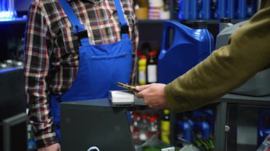 Bir otomotiv dükkanından motor yağı alırken bağlantısız bir terminalde akıllı telefonu dinleyerek satın almayı tamamlayan bir müşterinin yakın plan görüntüsü.