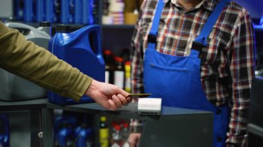 Bir otomotiv parçası mağazasından motor yağı alırken bir terminalde banka kartıyla temassız ödeme yapan müşterinin yakın çekimi.