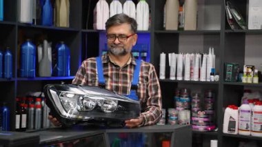 Gülümseyen kıdemli oto yedek parça satıcısı mavi tulumlu ve ekose gömlekli mağazadaki müşteriye yedek araba farı veriyor..
