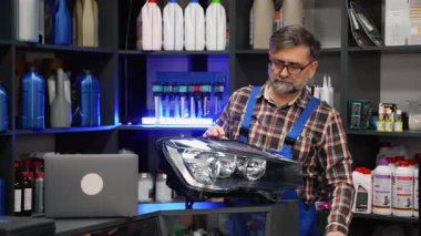 Tamirhanedeki kıdemli erkek satıcı, envanter kontrolü sırasında araba farını barkod okuyucusuyla tarıyor, dizüstü bilgisayarın yanında duruyor..