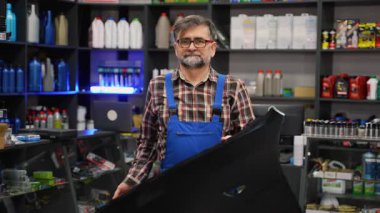 Ekose gömlek ve mavi tulumlu yaşlı erkek oto yedek parça satıcısı iyi stoklanmış bir otomotiv dükkanında dururken siyah bir araba tamponu tutuyor..