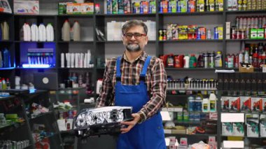 Gülümseyen yaşlı erkek oto yedek parça satıcısı, mavi tulumlu, iyi stoklanmış bir otomotiv deposunda parlak bir araba farına sahip..