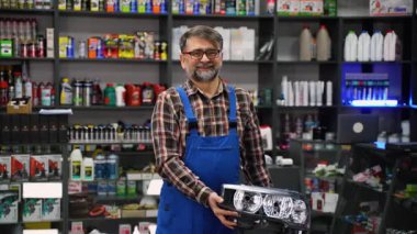 İyi stoklanmış bir otomotiv mağazasının içindeki müşteriye araba farı verirken mavi tulumlu arkadaş canlısı oto yedek parça satıcısı gülümsüyor..