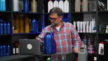 Kareli gömlekli yaşlı bir erkek, modern bir oto yedek parça mağazasında motor yağı stoklarını kontrol etmek için barkod tarayıcısı ve dizüstü bilgisayar kullanıyor..