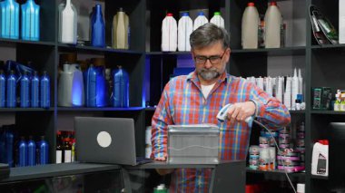 Yaşlı bir erkek çalışan araba aküsünü bir oto yedek parça dükkanında barkod okuyucusuyla tarıyor ve envanter sistemindeki stokları güncelliyor..