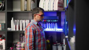 Ekose gömlekli yaşlı erkek müşteri, iyi stoklanmış bir oto yedek parça mağazasında parlak ışıkların altında, raftaki kamyon motor yağı şişelerini inceliyor..