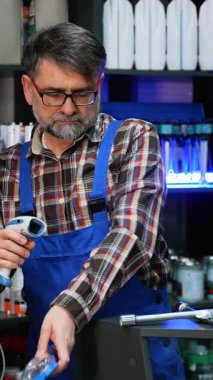 Ekose gömlek ve mavi tulumlu yaşlı bir erkek işçi, tezgahtaki metal bir otomotiv aletini barkod tarayıcısı kullanarak tarıyor..