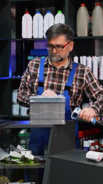 Ekose gömlekli ve tulumlu yaşlı erkek işçi araba aküsünde motor yağı rafları olan bir oto yedek parça mağazasında envanter taraması yapıyor..