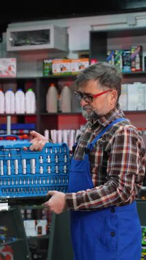 Ekose gömlekli ve tulumlu deneyimli bir erkek katip dolu bir oto yedek parça deposunda mavi bir anahtar takıyor ve sunuyor..