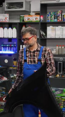 Ekose gömlek ve mavi tulumlu yaşlı erkek oto yedek parça satıcısı iyi stoklanmış bir otomotiv dükkanında dururken siyah bir araba tamponu tutuyor..