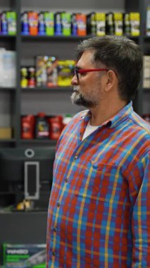 Renkli tişörtlü yaşlı bir adam bir otomotiv mağazasında motor yağı raflarını inceler ve aracı için doğru ürünü dikkatle seçer..