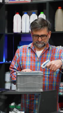 Yaşlı bir erkek çalışan araba aküsünü bir oto yedek parça dükkanında barkod okuyucusuyla tarıyor ve envanter sistemindeki stokları güncelliyor..