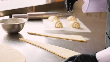 Eldivenli fırıncı, profesyonel mutfakta taze kruvasan hazırlarken metal masadaki üçgen şekilli hamur parçalarının üzerine un tozu sürer..