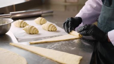 Profesyonel şef dikkatli bir şekilde üçgen hamuru kruvasana çeviriyor. Yanında hamur işleri olan bir fırın mutfağında un metal bir yüzey üzerinde..