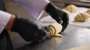 Bir fırın mutfağında, paslanmaz çelik bir yüzey üzerinde kruvasan hamurunun rulosunu dikkatlice bitiren, eldivenli şeflerin yakın çekimi..