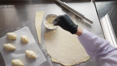 Çiğ kruvasanlar parşömen üzerinde, aletlerle ve un kasesiyle paslanmaz tezgahta dinlenirken, fırıncının un tozu hamurcuğun üzerinde..