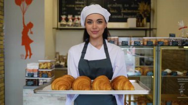 Şef üniformalı genç bir kadın sıcak bir pastanenin içinde gururla gülümserken elinde bir tepsi dolusu yeni pişmiş kruvasan tutuyor..