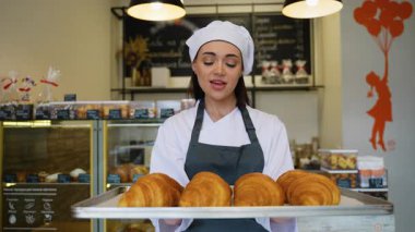 Şef üniformalı oyuncu kadın, sıcak bir pastanede taze pişmiş altın kruvasan sunarken sevinçle şarkı söylüyor..