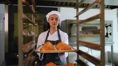 Fırın üniformalı kadın dikkatlice bir tepsi taze pişmiş kruvasanı ticari bir mutfaktaki metal soğutma rafına koyuyor..