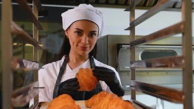 Aşçı üniformalı kadın fırıncı iki elinde altın bir kruvasan tutarken sıcak bir şekilde gülümsüyor, profesyonel bir mutfakta soğutma rafının yanında duruyor..