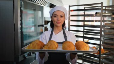 Üniformalı genç bayan şef, arkasında soğutucu rafları olan ticari bir mutfakta bir tepsi altın kruvasanı gururla sunar..