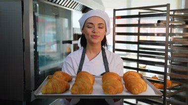 Üniformalı kadın aşçı, elinde bir tepsi taze kruvasan tutarken gözleri kapalı gülümser ve fırın mutfağındaki lezzetli aromanın tadını çıkarır..