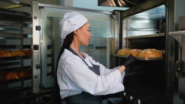 Üniformalı profesyonel kadın fırıncı bir fırının mutfağından bir tepsi altın, kabuklu ekmek alıyor..