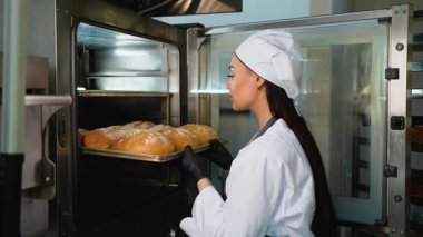 Aşçı üniformalı kadın profesyonel bir fırın ortamında ticari bir fırından bir tepsi taze, altın somun ekmek alıyor..