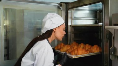 Üniformalı ve eldivenli kadın fırıncı, fırındaki bir tepsi dolusu yeni pişmiş kruvasanı dikkatlice çıkarıyor..
