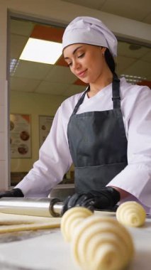 Aşçı üniformalı genç bir kadın, pencerenin yakınındaki temiz bir pastanede kruvasan hazırlarken hamura merdane takıyor..