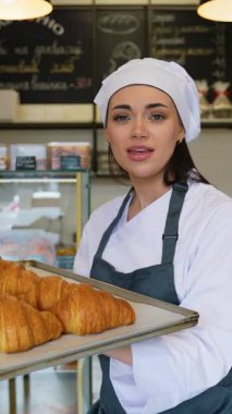 Şef üniformalı neşeli kadın, sıcak bir pastanede elinde taze pişmiş altın kruvasan tepsisiyle neşe içinde şarkı söyleyip dans ediyor..